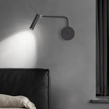 "A black wall lamp with a pivoting arm and cylindrical head, lit up on a gray wall, next to a black bed and curtains."