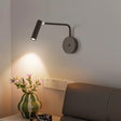 "A black wall lamp with a pivoting arm and cylindrical head, lit up on a gray wall, above a bedside table with flowers and next to a gray headboard."