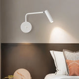 "A white wall lamp with a pivoting arm and cylindrical head, installed above a bedside table with a round object, next to a dark brown headboard."