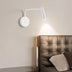 "A white wall lamp with a pivoting arm and cylindrical head, installed above a bedside table, next to a brown leather headboard."