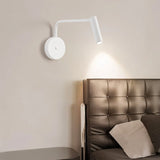 "A white wall lamp with a pivoting arm and cylindrical head, installed above a bedside table, next to a brown leather headboard."