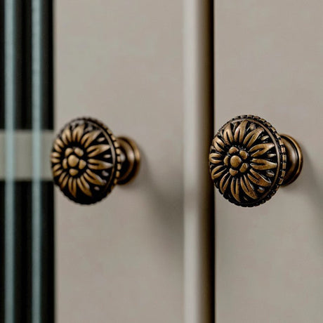 "Two antique brass floral knobs with raised petal detailing on a light gray cabinet."
