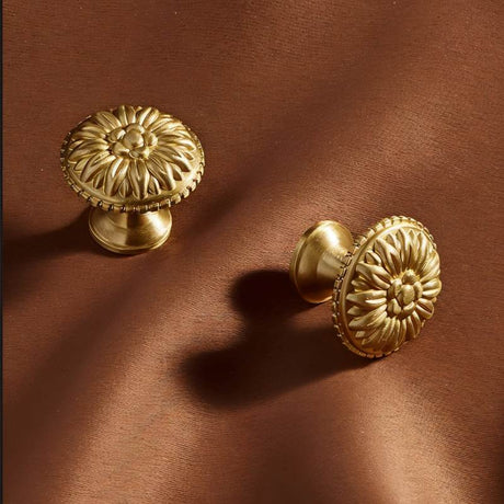 "Two gold brass floral knobs with raised petal detailing on a brown fabric."