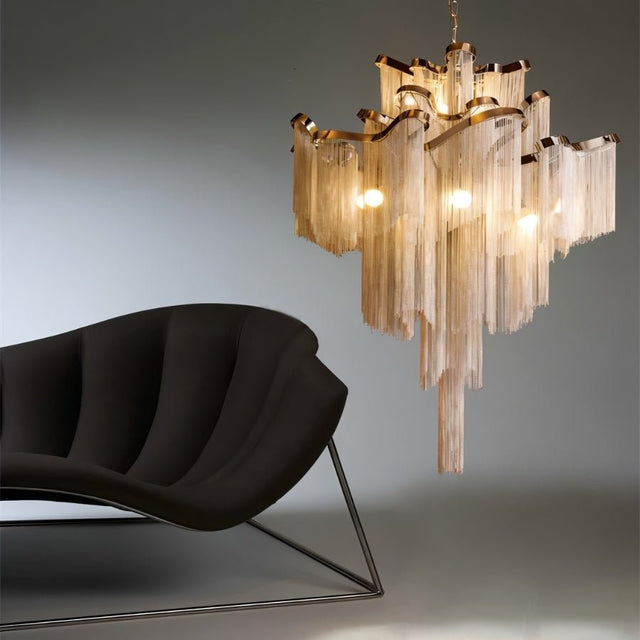 Nazra Chandelier – Open Box in gold finish with cascading metallic fringe, paired with modern black lounge chair for luxury living room lighting.