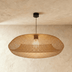 Woven wicker pendant light with curved organic shape in natural tan finish, suspended in modern bedroom above bed with pillow