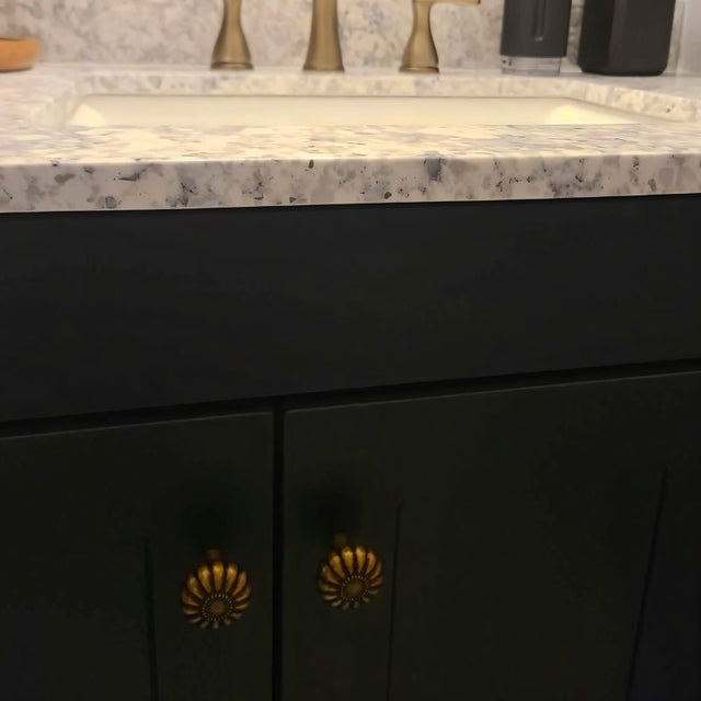 "Two vintage brass flower-shaped knobs with bold, rounded petals, installed on dark green bathroom cabinets, complementing the white countertop and sink."