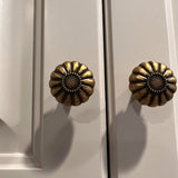 "Two vintage brass flower-shaped knobs with bold, rounded petals, installed on a gray cabinet, showcasing their intricate embossed pattern and antique appeal."
