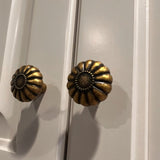 "Two vintage brass flower-shaped knobs with bold, rounded petals, installed on a gray cabinet, showcasing their intricate embossed pattern and antique appeal."