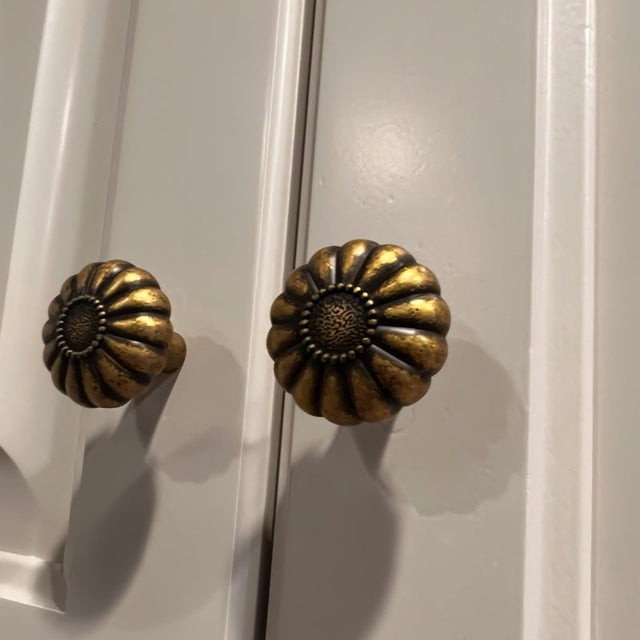 "Two vintage brass flower-shaped knobs with bold, rounded petals, installed on a gray cabinet, showcasing their intricate embossed pattern and antique appeal."