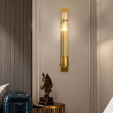 "A modern brass wall lamp with a rectangular base and a cylindrical glass shade mounted on a light-colored wall in a hallway, illuminating a horse head sculpture on a side table."