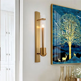 "A modern brass wall lamp with a rectangular base and a cylindrical glass shade mounted on a white wall in a living room, next to a framed painting of a tree."