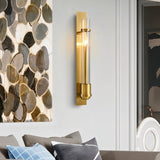 "A modern brass wall lamp with a rectangular base and a cylindrical glass shade illuminates a contemporary living room with abstract art on the wall."