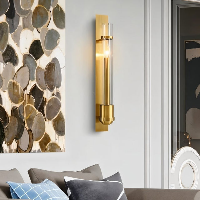 "A modern brass wall lamp with a rectangular base and a cylindrical glass shade illuminates a contemporary living room with abstract art on the wall."