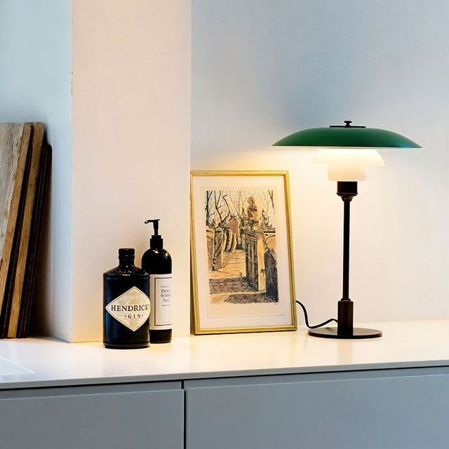 "Nisra Table Lamp with a green top shade and frosted glass diffuser, placed on a surface next to a framed artwork and bottles, made from aluminum, iron, and glass for soft, ambient lighting."