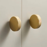 "Two gold brass cylindrical knobs with knurled texture on a light gray cabinet door."