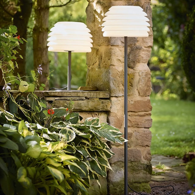 "A white table lamp with a layered, wave-like diffuser, sitting on a stone ledge in a garden next to a matching floor lamp."