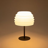 "A white table lamp with a layered, wave-like diffuser, lit up with a warm glow against a warm-toned background."