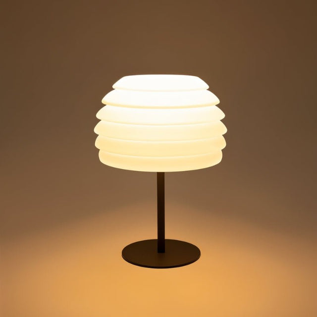 "A white table lamp with a layered, wave-like diffuser, lit up with a warm glow against a warm-toned background."