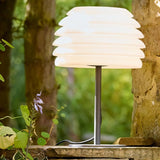 "A white table lamp with a layered, wave-like diffuser, lit up on a stone ledge in a garden with plants."