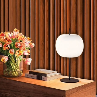 "Modern table lamp with frosted glass shade, aluminum and iron base, placed next to colorful flowers, creating a warm, inviting glow in a contemporary living room setting."