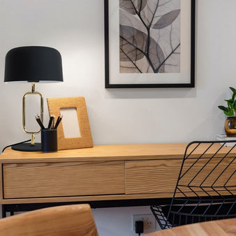 "A modern black table lamp with a brass oval frame sits on a light wood desk in a contemporary office setting with a framed picture and a wire chair."