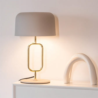 "A modern grey table lamp with a brass oval frame and a dome shade, placed on a white surface next to a decorative arched object."