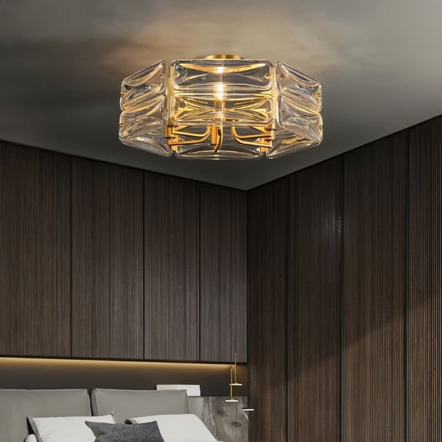"A modern flush mount ceiling light with a geometric glass shade and gold frame, installed above a bed in a bedroom with wood paneling."