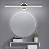 "A modern gray wall lamp with a cylindrical bar intersecting a circular base, mounted above a round mirror in a contemporary bathroom with a gray tiled wall and a small shelf with towels."