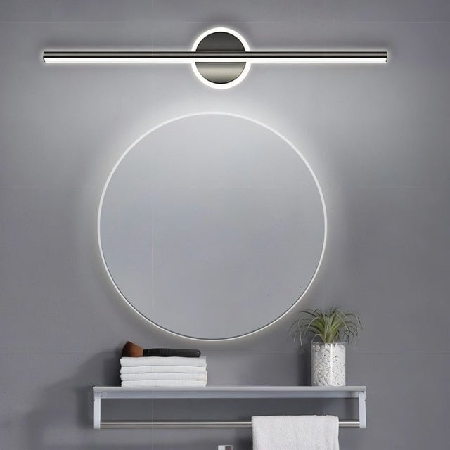 "A modern gray wall lamp with a cylindrical bar intersecting a circular base, mounted above a round mirror in a contemporary bathroom with a gray tiled wall and a small shelf with towels."