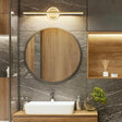 "A modern gold wall lamp with a cylindrical bar intersecting a circular base, mounted above a round mirror in a contemporary bathroom with gray tiled walls."