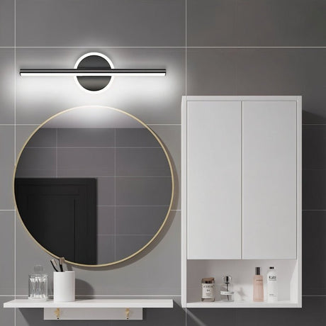 "A modern black wall lamp with a cylindrical bar intersecting a circular base, mounted above a round mirror in a contemporary bathroom with gray tiled walls."