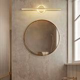 "A modern gold wall lamp with a cylindrical bar intersecting a circular base, mounted above a round mirror in a contemporary bathroom with textured walls."
