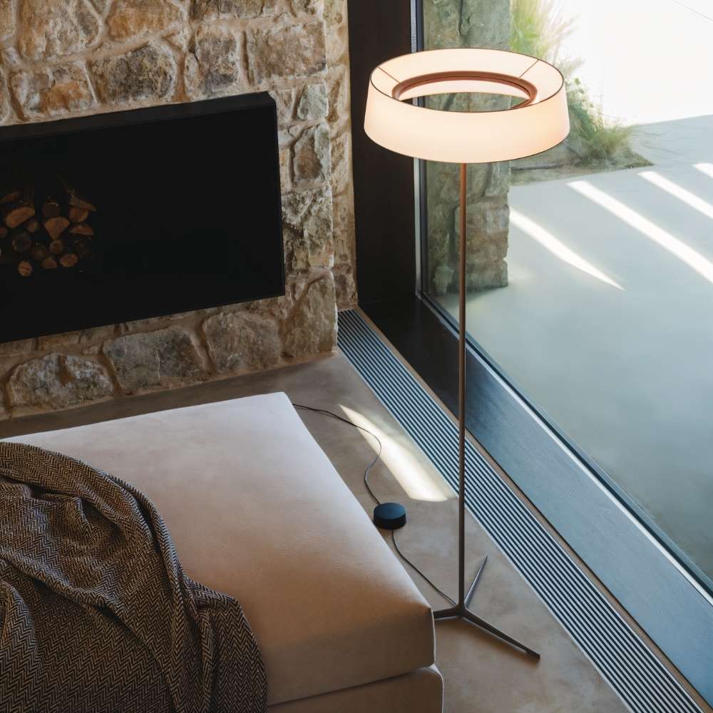 "A contemporary floor lamp with a ring-shaped fabric shade stands next to a large window in a modern living room with stone walls."