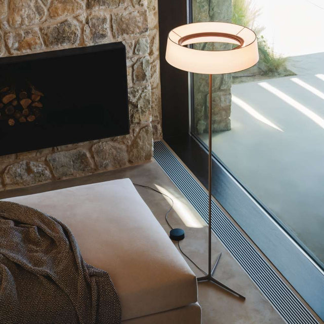 "A contemporary floor lamp with a ring-shaped fabric shade stands next to a large window in a modern living room with stone walls."