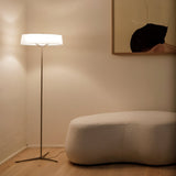 "A minimalist floor lamp with a rectangular fabric shade and a slim stand sits in a corner of a living room with a white sofa and artwork on the wall."