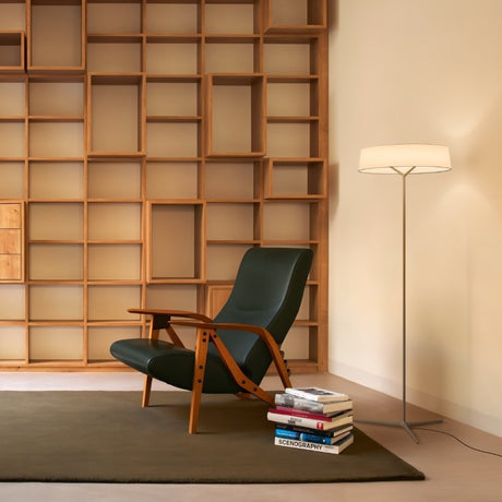 "A floor lamp with a ring-shaped fabric shade illuminates a reading nook with a modern chair, a stack of books titled 'Scenography', and a large, compartmentalized bookshelf."