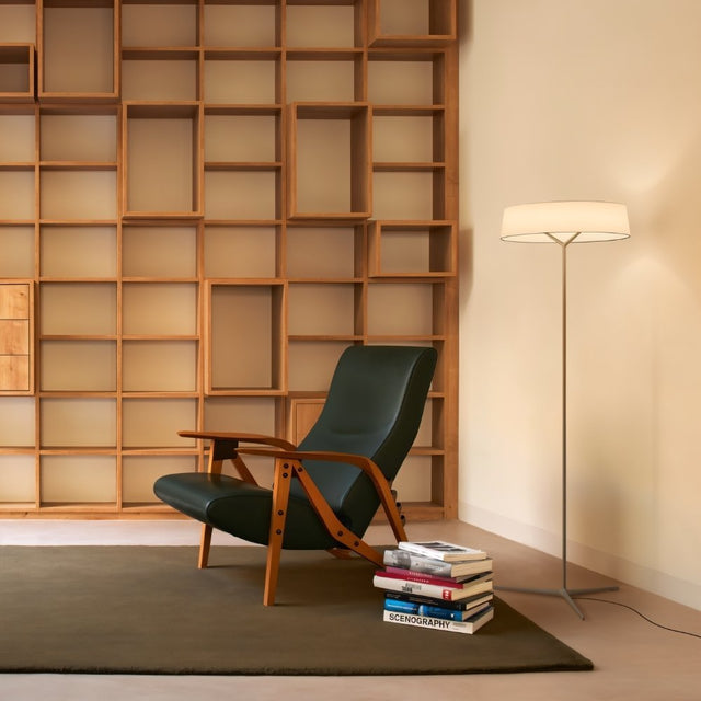 "A floor lamp with a ring-shaped fabric shade illuminates a reading nook with a modern chair, a stack of books titled 'Scenography', and a large, compartmentalized bookshelf."