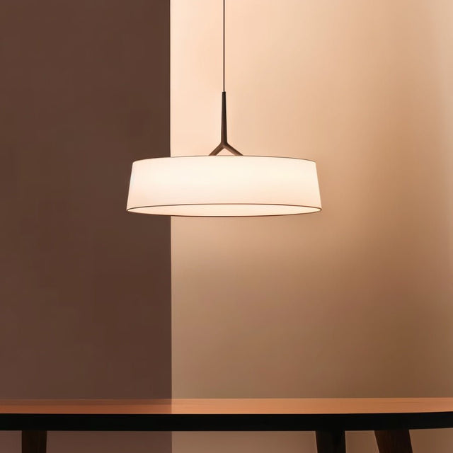 "A pendant light with a wide, white fabric drum shade and a dark frame hangs in a room with warm, neutral tones."