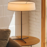 "A contemporary table lamp with a fabric drum shade sits on a round side table next to a sofa in a modern living room."
