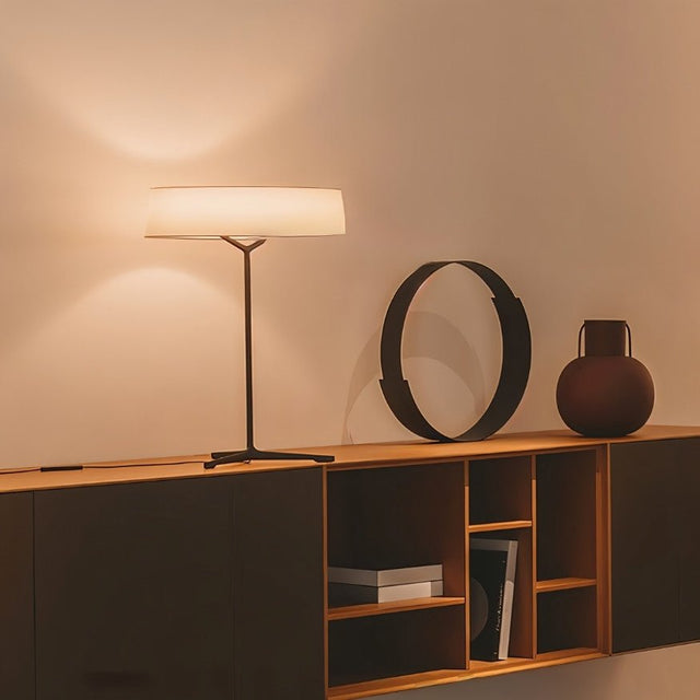 "A modern table lamp with a fabric drum shade sits on a dresser in a dimly lit bedroom, providing a soft glow."