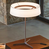"A modern table lamp with a white fabric drum shade and a sleek, light-colored stand sits on a wooden nightstand in a bedroom."
