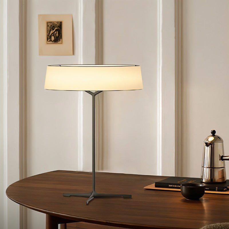 "A modern table lamp with a fabric drum shade sits on a round wooden table in a well-lit living room."