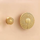 "Two gold brass knobs with geometric lattice designs, in different sizes, mounted on a light pink surface."