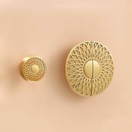 "Two gold brass knobs with geometric lattice designs, in different sizes, mounted on a light pink surface."