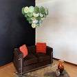 "A modern, round chandelier with cascading green and iridescent glass discs, hanging in an eclectic living room with a brown sofa and colorful accents."