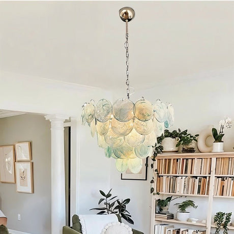 "A modern, round chandelier with cascading blue and iridescent glass discs, hanging in a bright living room with plants and a bookshelf."