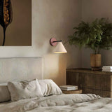 "A modern wall lamp with a conical travertine shade and a black arm, mounted on a wooden backplate in a bedroom, providing a soft, warm glow."
