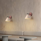 "A pair of modern wall lamps with conical travertine shades and black arms, mounted on a textured wall, providing a soft, warm glow above a seating area."