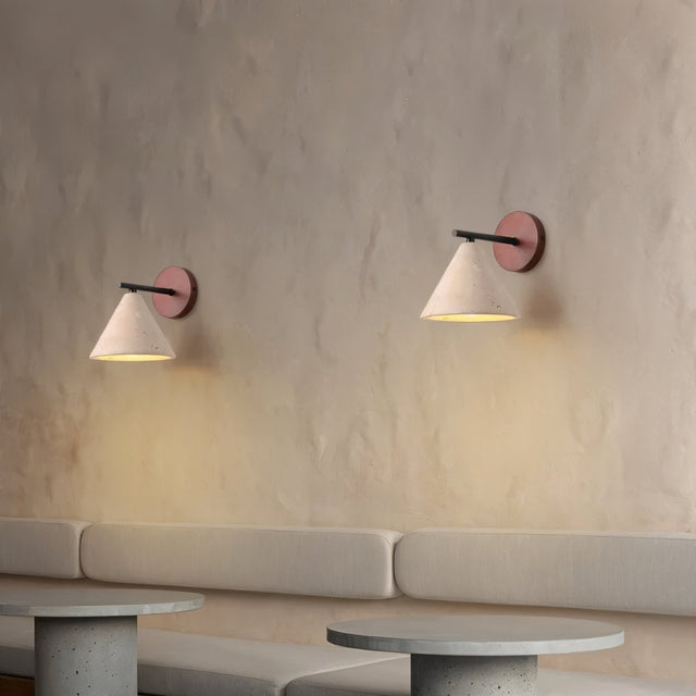 "A pair of modern wall lamps with conical travertine shades and black arms, mounted on a textured wall, providing a soft, warm glow above a seating area."