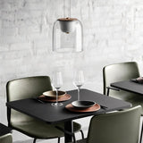 "A modern pendant light with a clear glass shade and wooden top, hanging above a dining table with place settings and wine glasses."